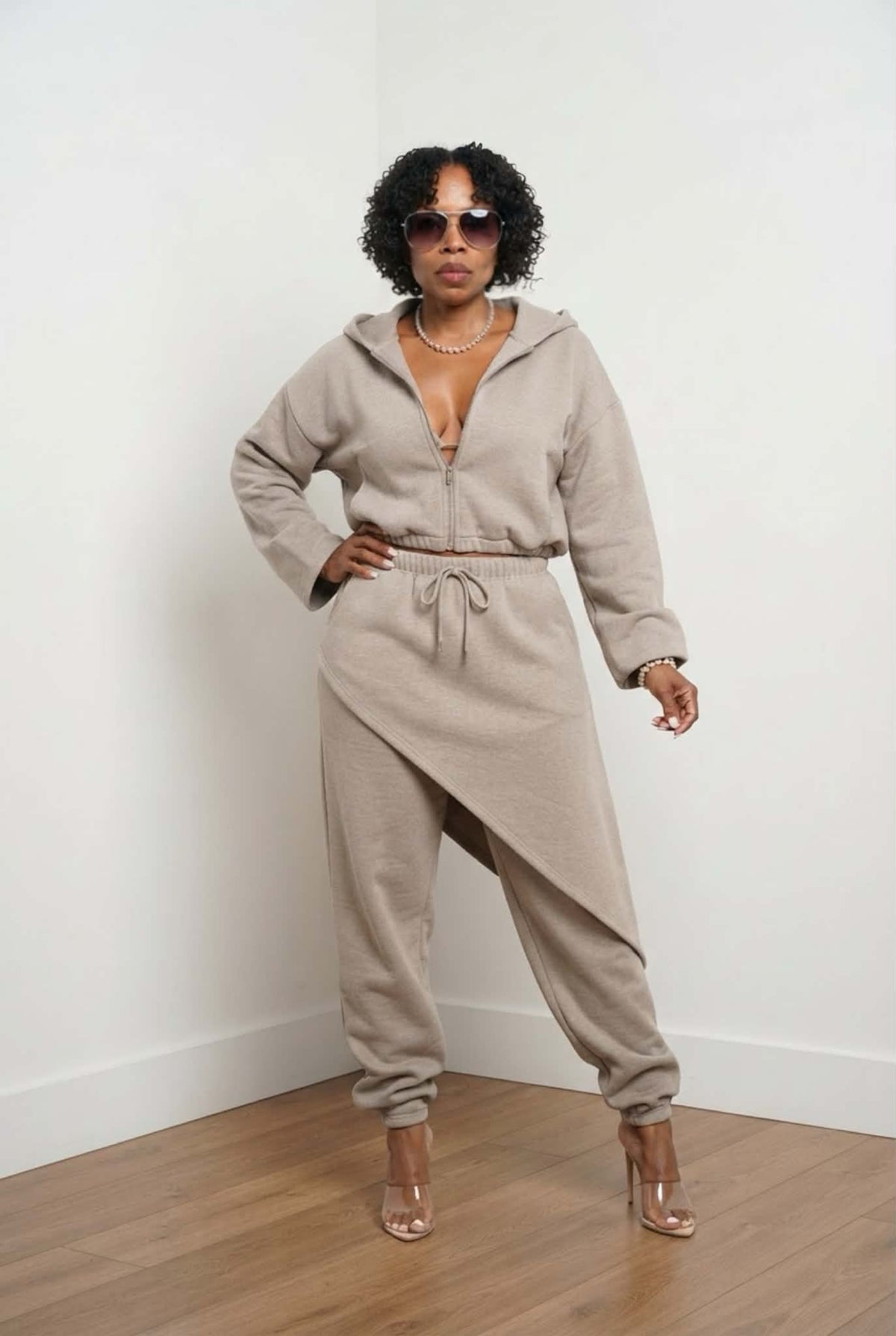 Beige two-piece lounge set featuring a cropped zip-up hoodie and asymmetrical wrap-detail jogger pants, styled with oversized sunglasses and clear heels in a studio setting.