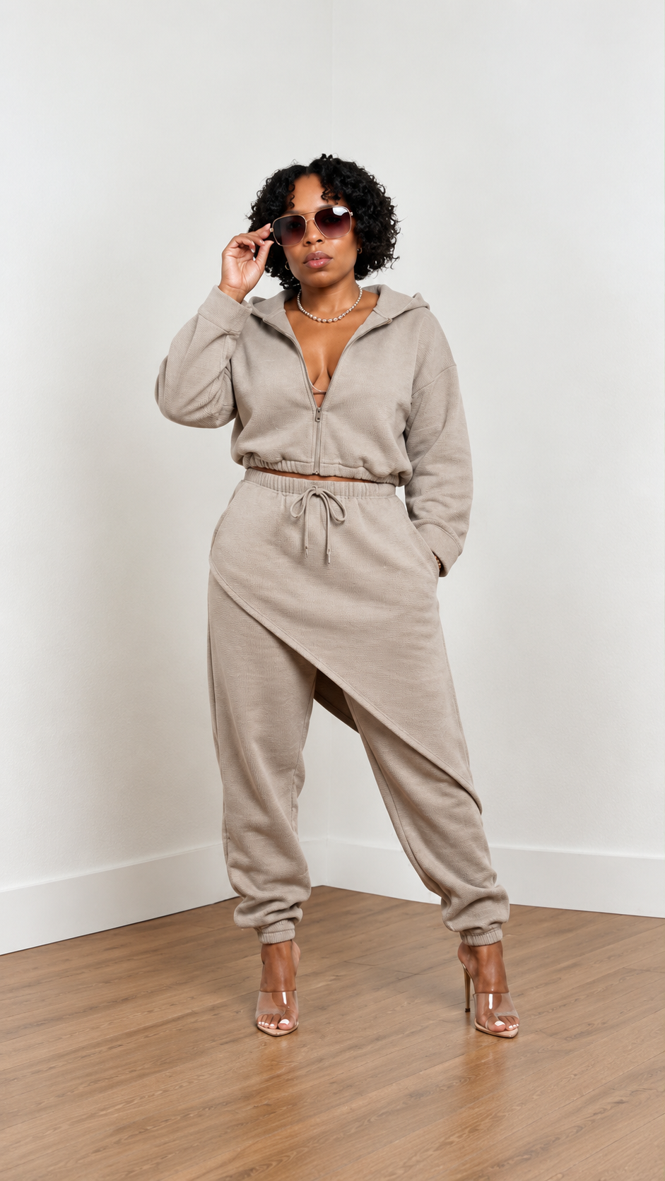 Beige two-piece lounge set featuring a cropped zip-up hoodie and asymmetrical wrap-detail jogger pants, styled with oversized sunglasses and clear heels in a studio setting.